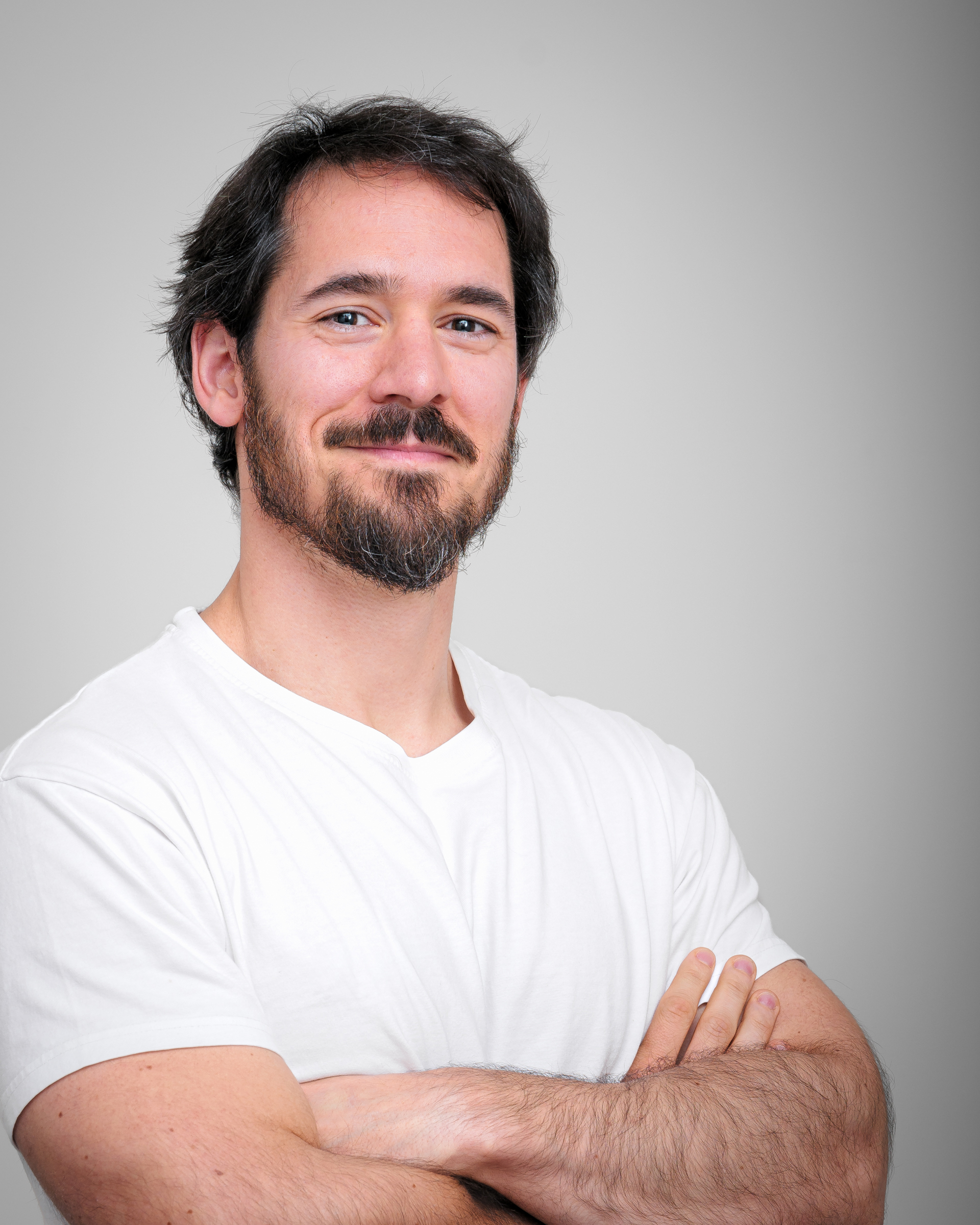 A light-skinned man in his mid-30s with dark brown, slightly wavy hair and a well-groomed beard stands against a neutral gray background. His eyes are deep-set and dark, and he has a confident, warm smile. He is wearing a plain white T-shirt and has a relaxed posture with his arms crossed. His muscular arms and lightly tanned skin suggest an active lifestyle. His expression is approachable and self-assured, conveying a sense of quiet confidence (AI generated).